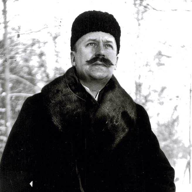 Black-and-white portrait of a Harald Pihl in a fur coat and hat, standing outdoors in winter.